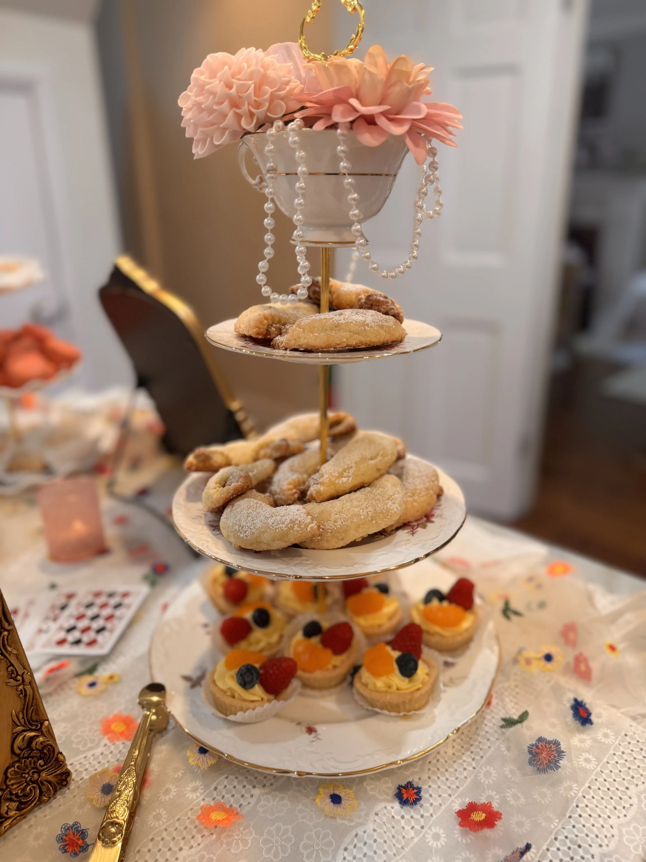 A three-tiered serving stand filled with assorted cookies and small tarts, decorated with pink artificial flowers and pearl-like beads, set on a floral embroidered tablecloth.