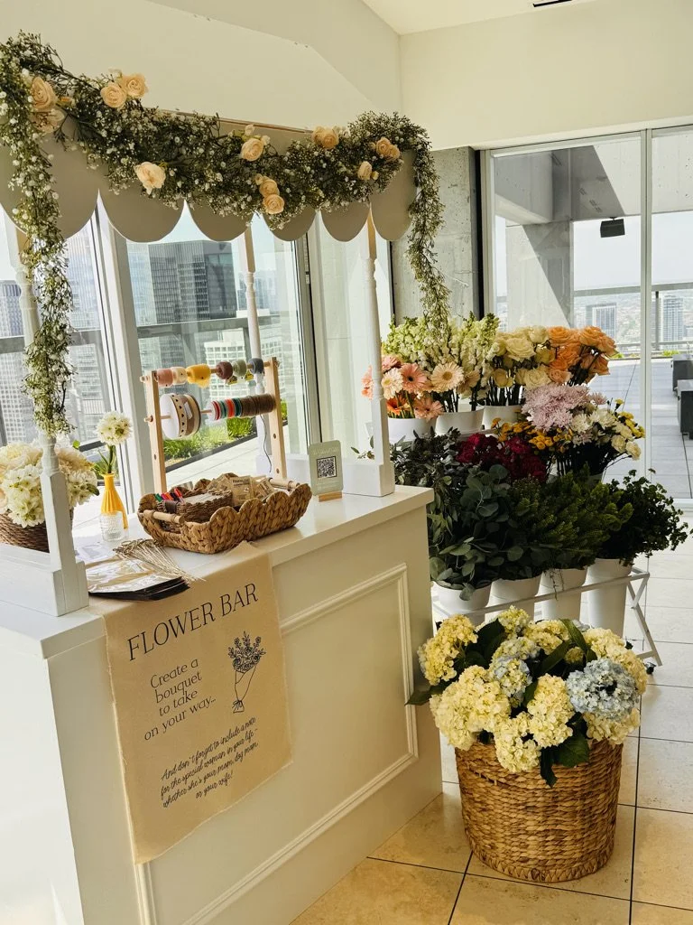 Flower stand with various bouquets and a flower-themed 'Flower Bar' sign, located in a bright indoor space with large windows.