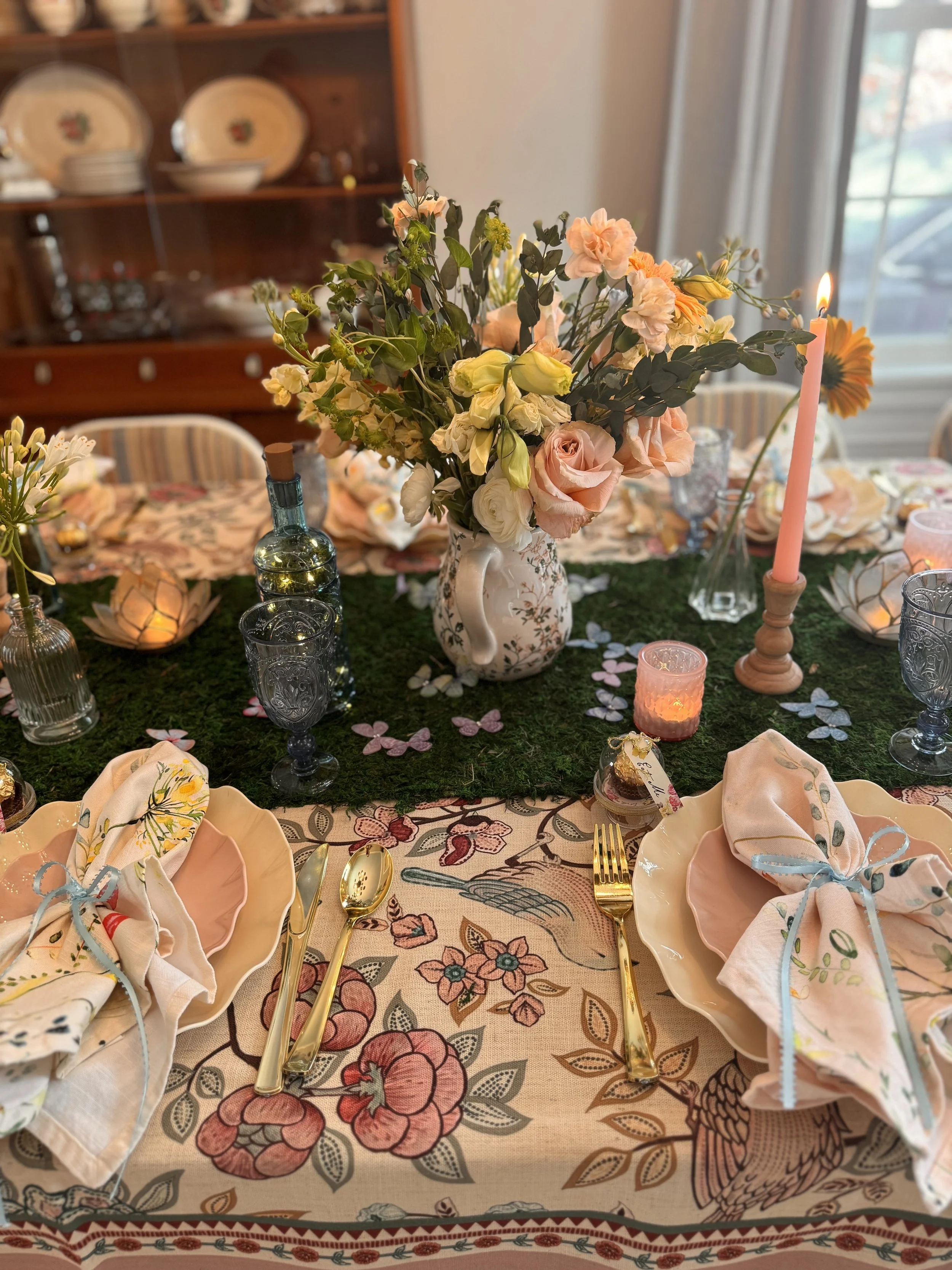 A table setting with a floral centerpiece, pink and white napkins with blue ribbons, gold utensils, and candles. The tablecloth features a floral and bird pattern. There are decorative items and an interior background with a china cabinet and windows.