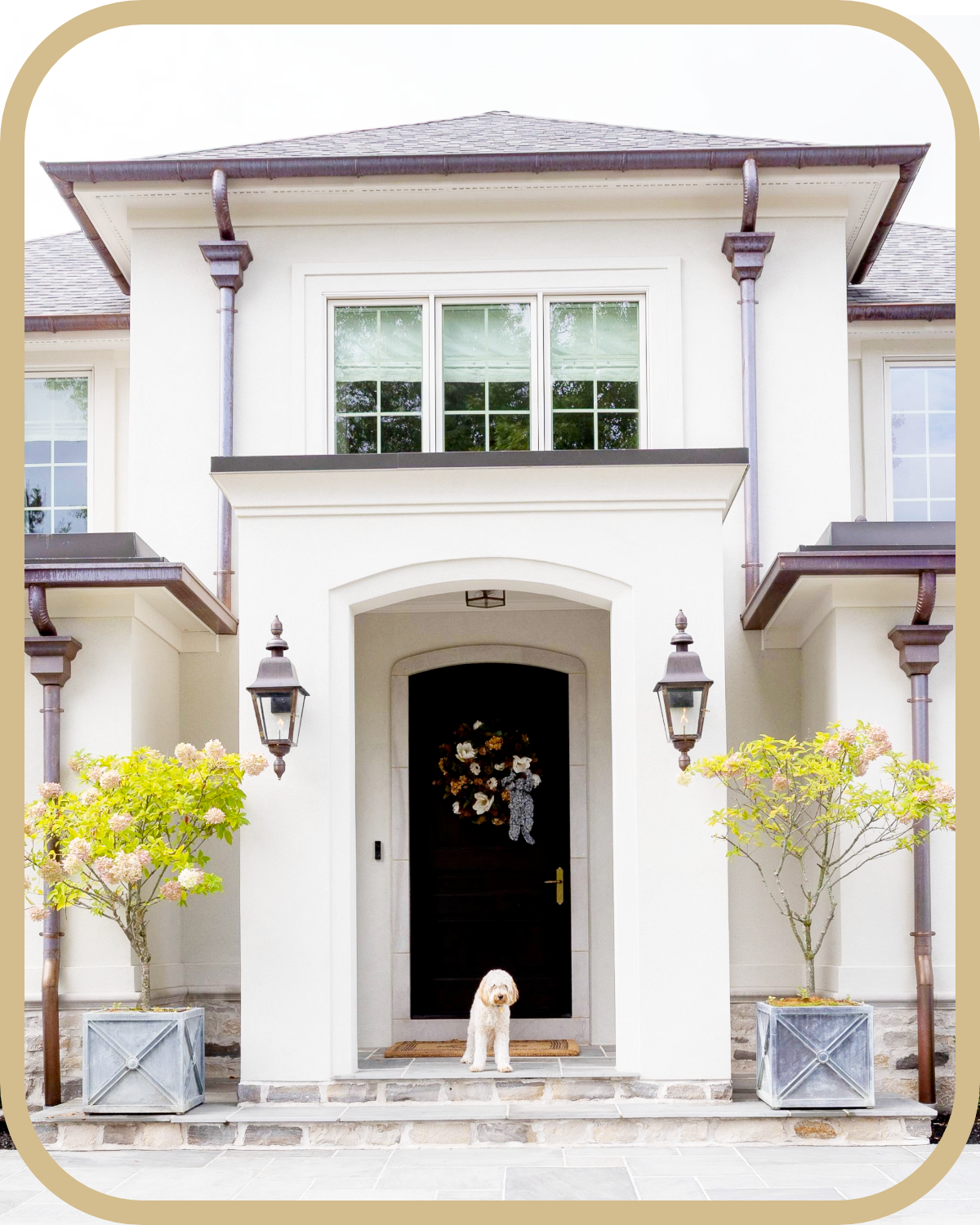 Front entrance of a white two-story stucco house with a black door, two outdoor lanterns, potted trees, a small white dog sitting in front, and a floral wreath on the door.