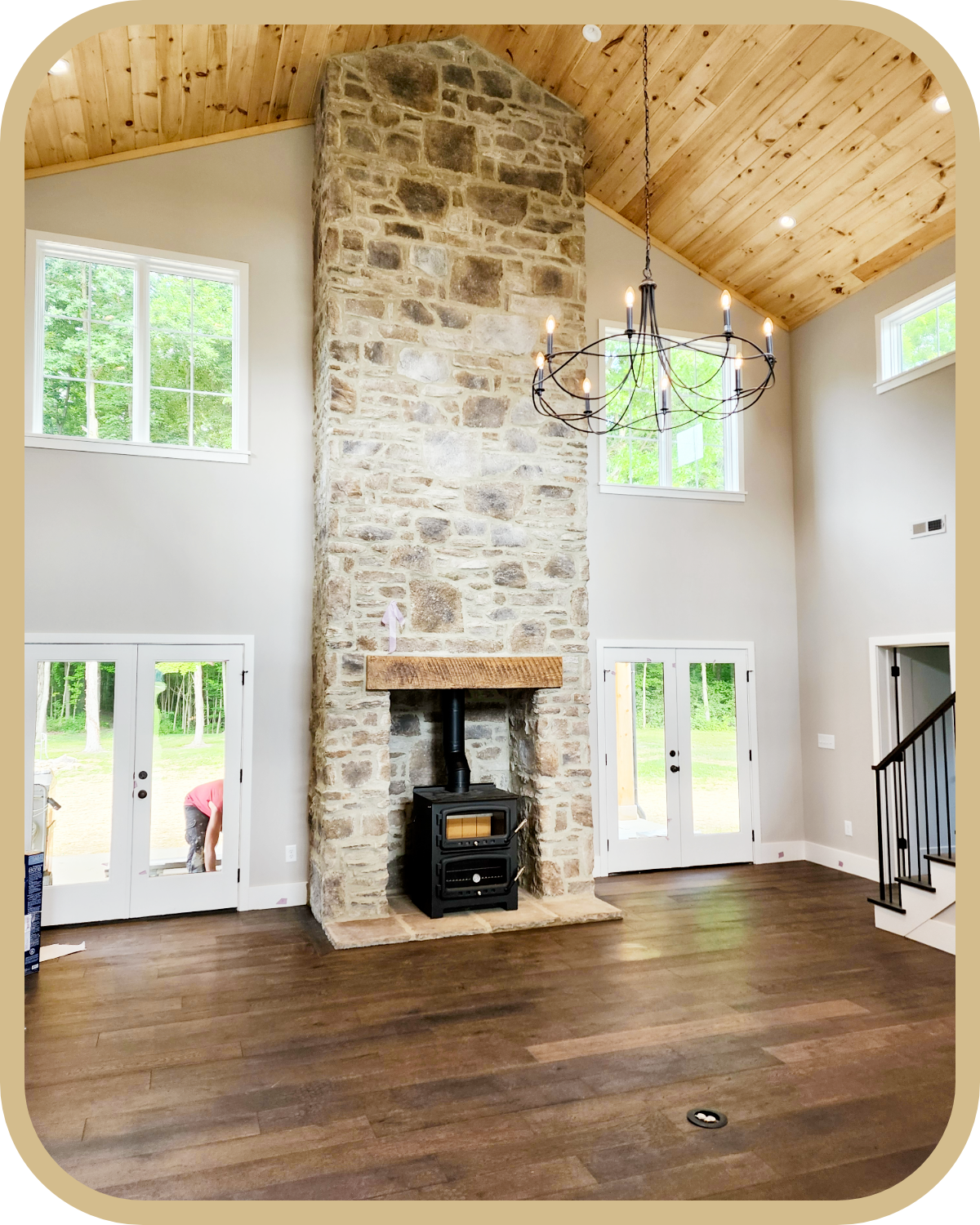 Interior of a living room with a tall stone fireplace, a wooden mantel, hardwood flooring, and large windows. A black stove is in the fireplace, and a chandelier hangs from the wooden ceiling. Double glass doors lead outside, revealing a green outdoor scene.