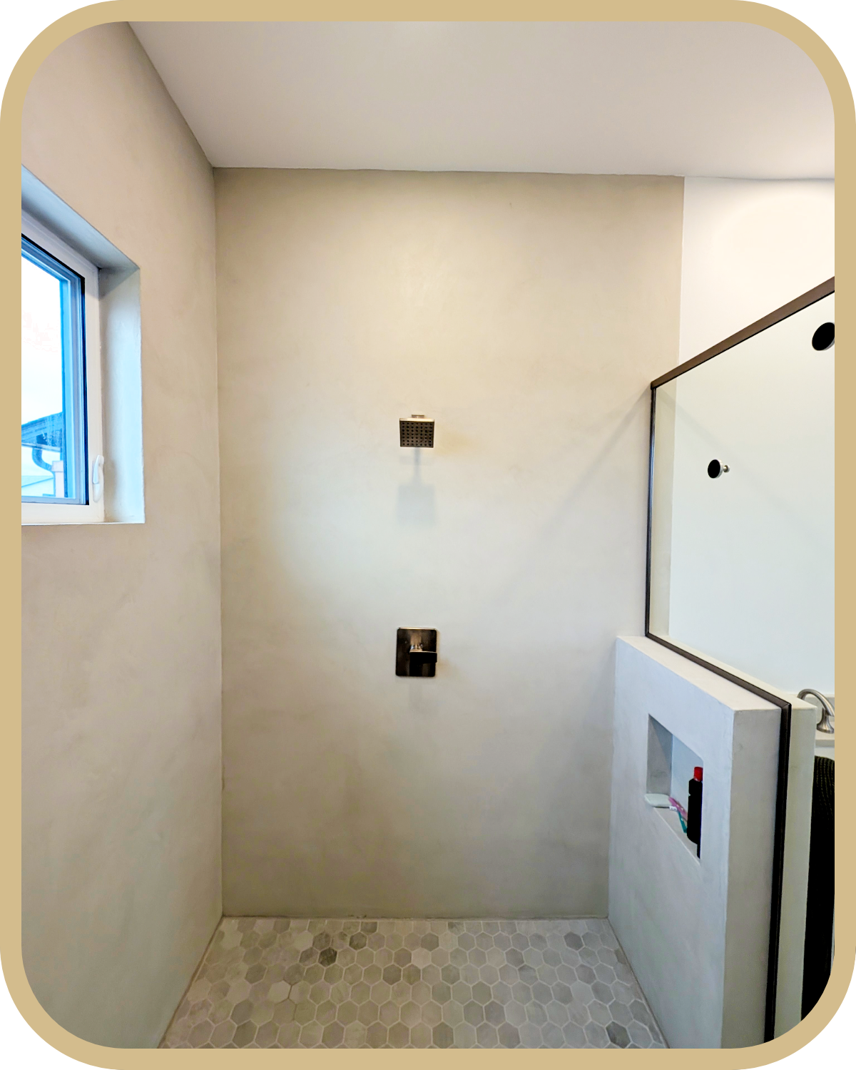 A modern bathroom shower area with Microcement applied on the walls and a small window, hexagonal floor tiles, a ceiling shower head, and a built-in shelf in the wall.