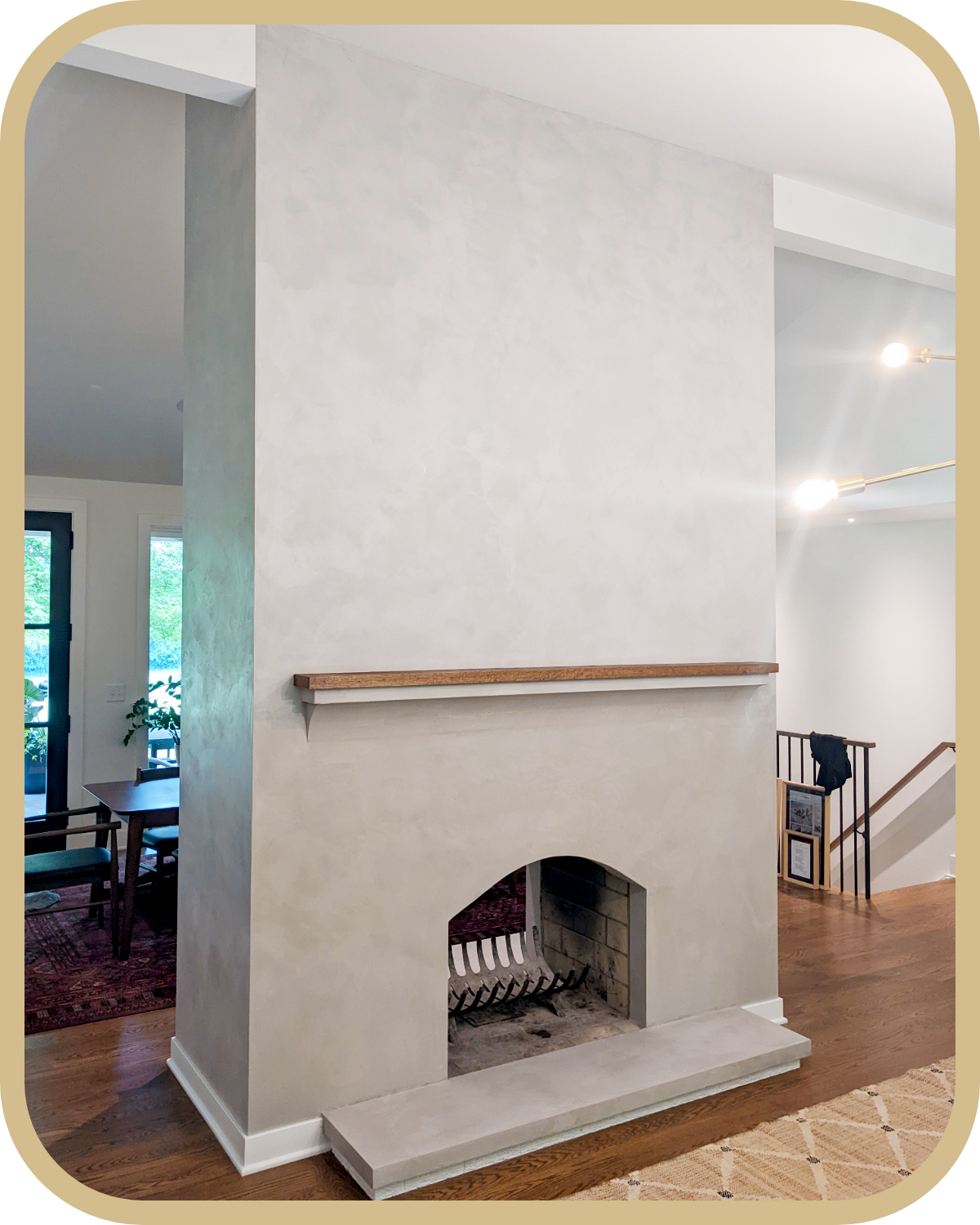 Living room fireplace with a beige plaster finish, a wooden mantel, and a fire grate inside. Part of a dining area and staircase are visible in a modern home interior.