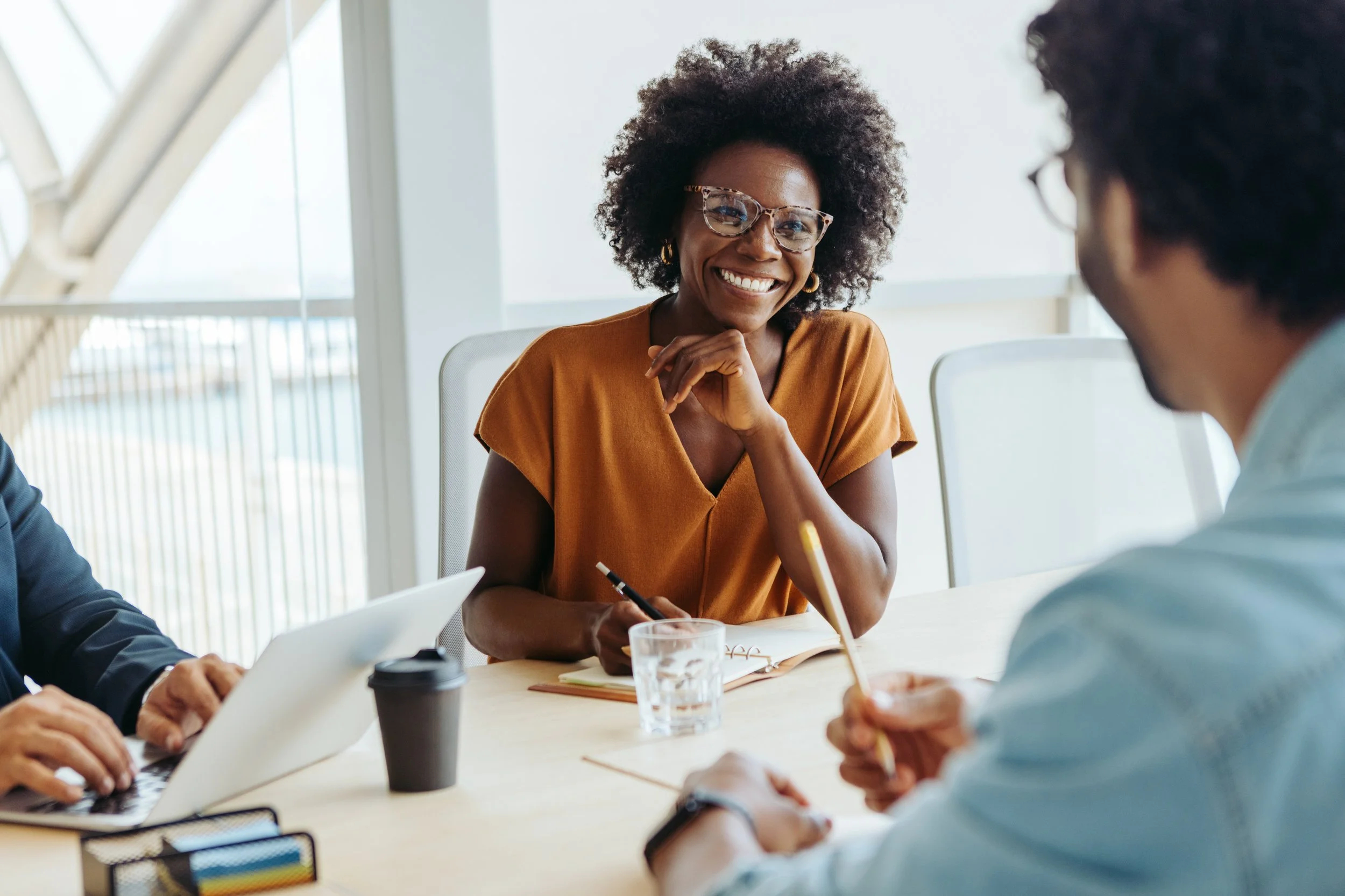 A diverse group of three people having a business meeting in a modern office, smiling and engaging in conversation.