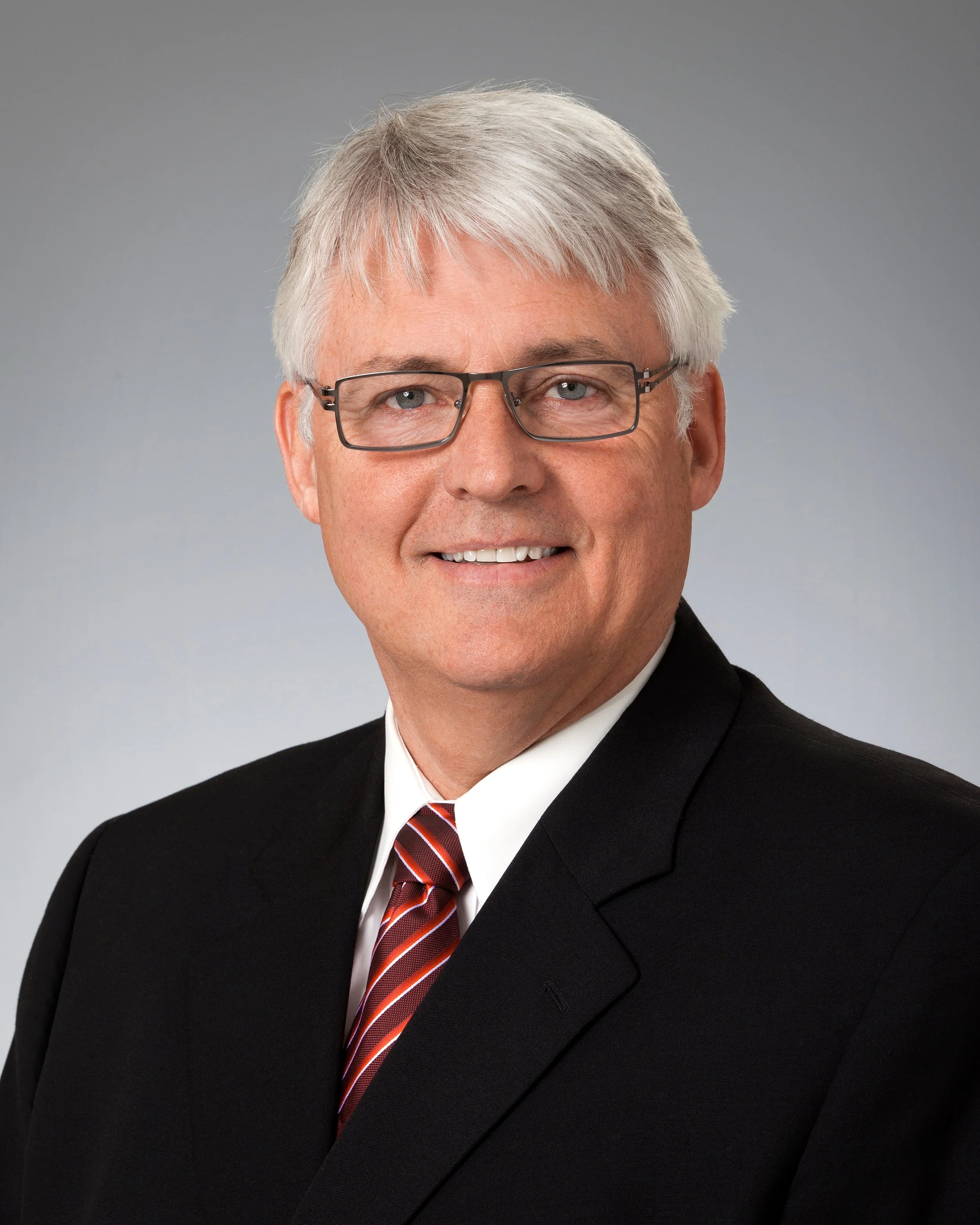 Professional headshot of a middle-aged man with gray hair, wearing glasses, a black suit, white shirt, and a red striped tie, smiling against a plain gray background.