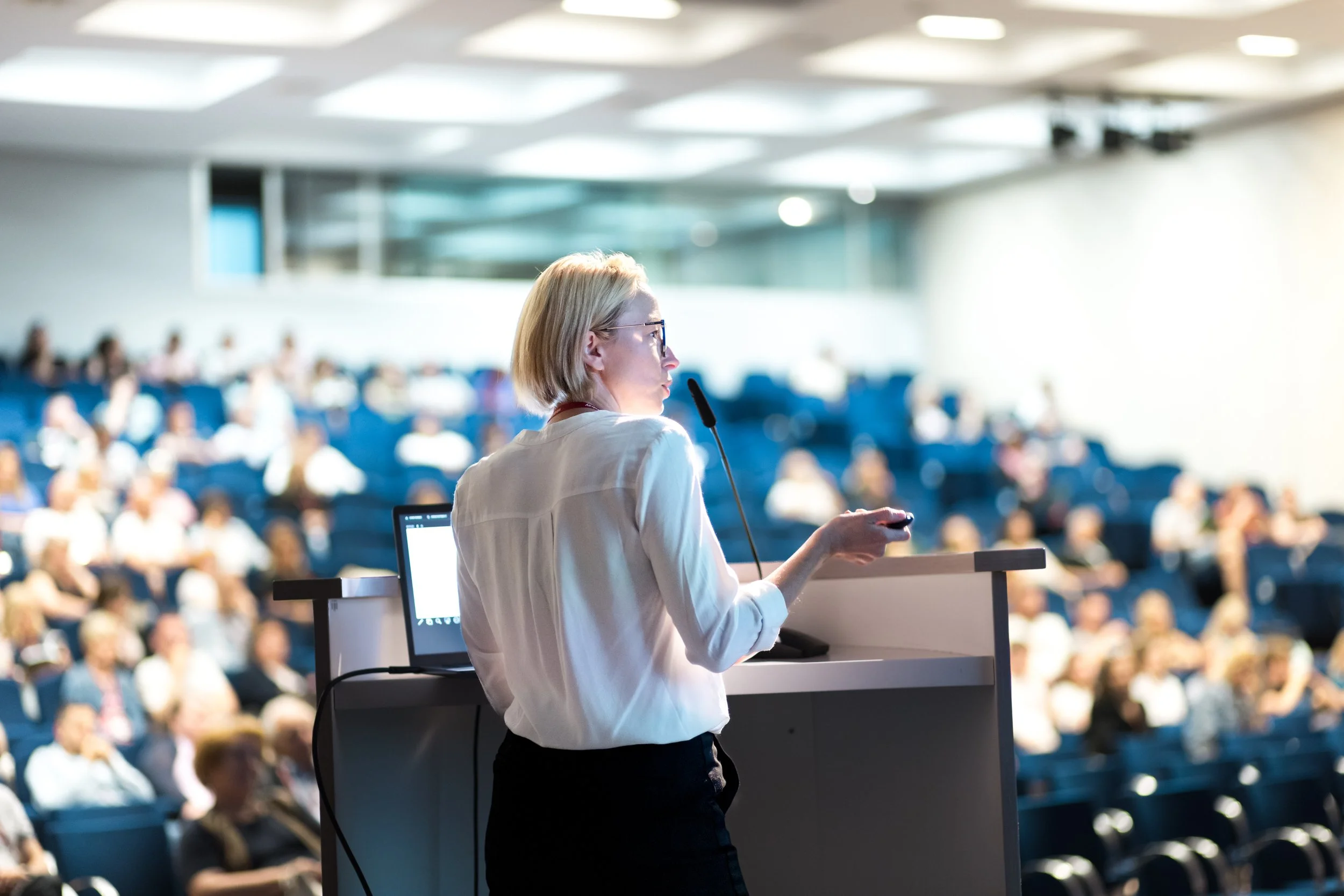 A woman with blonde hair and glasses giving a presentation at a conference in a large auditorium filled with seated attendees.