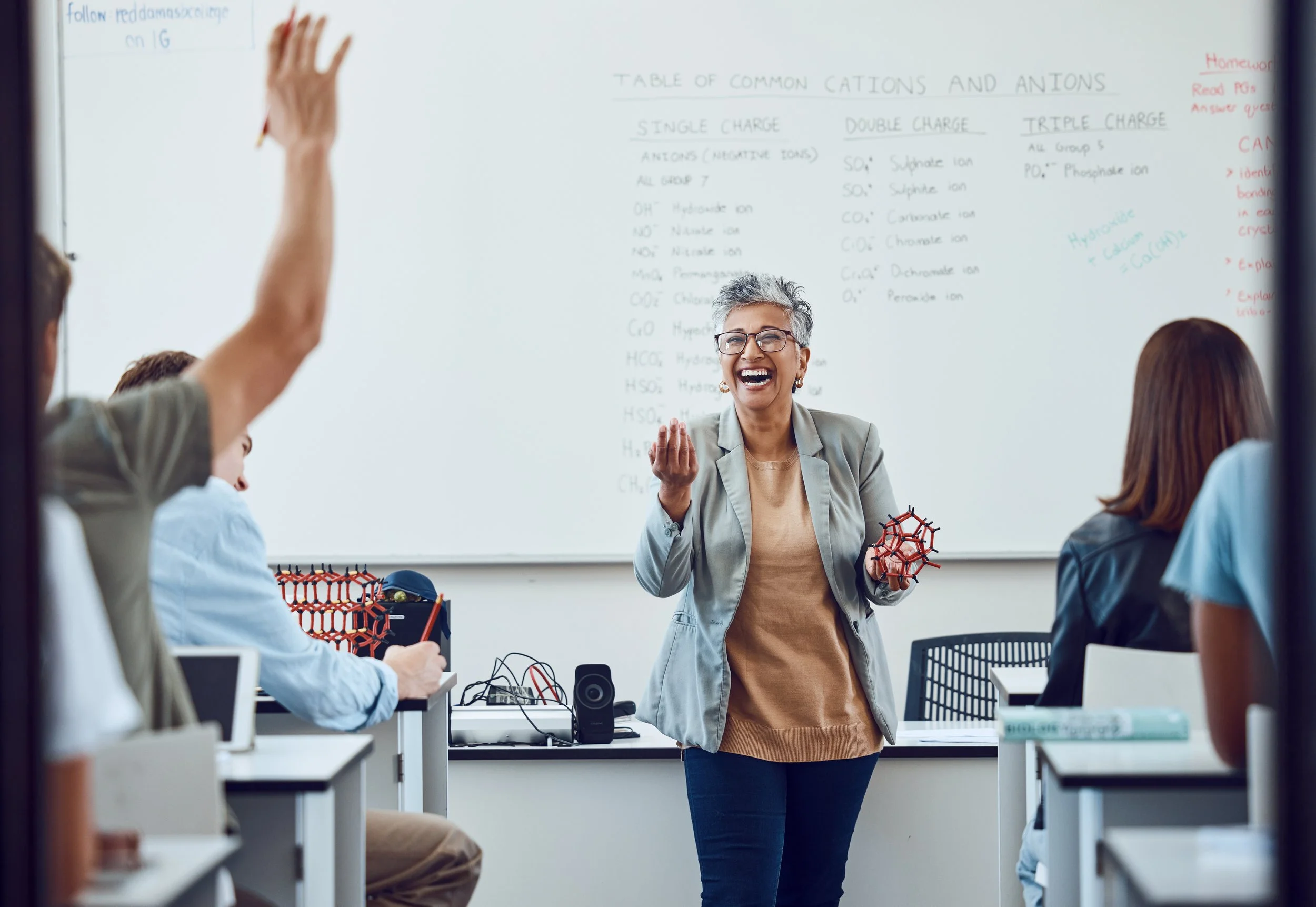 A teacher with short gray hair, glasses, and a beige shirt laughs while holding molecular models in a classroom. Students sit at desks, some raising hands, with a whiteboard behind her displaying a chart on chemical cations and anions.