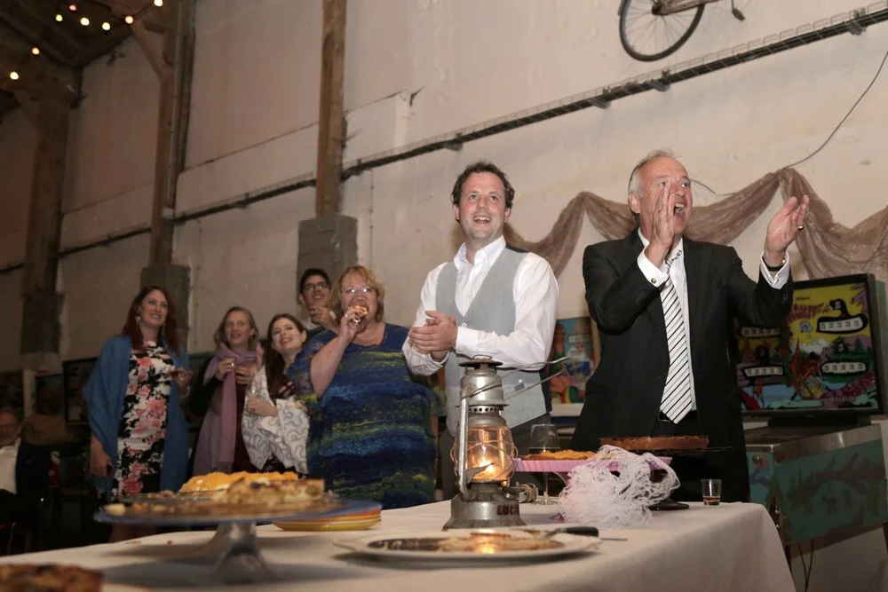 A group of people celebrating at a party in an industrial-style venue, with a man in a suit applauding near a table with food, and others standing, watching and smiling in the background.