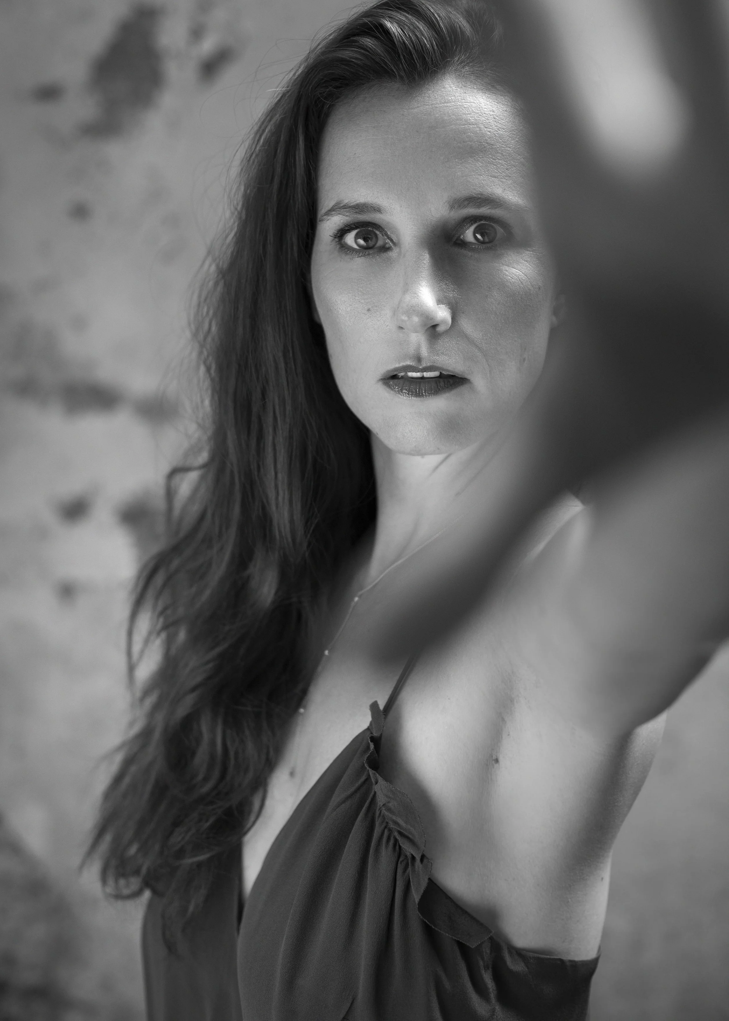 A black-and-white close-up portrait of a woman with long, wavy hair, and intense facial expression, wearing a sleeveless dress with a necklace, reaching towards the camera.