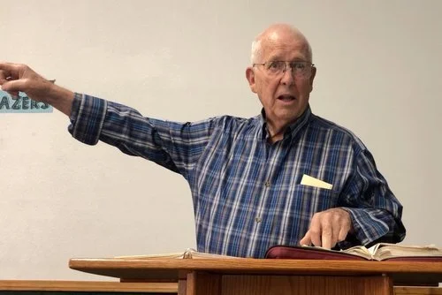 An elderly man with glasses, wearing a blue plaid shirt, standing at a podium in a classroom, gesturing with his right arm extended, with an open book in front of him.