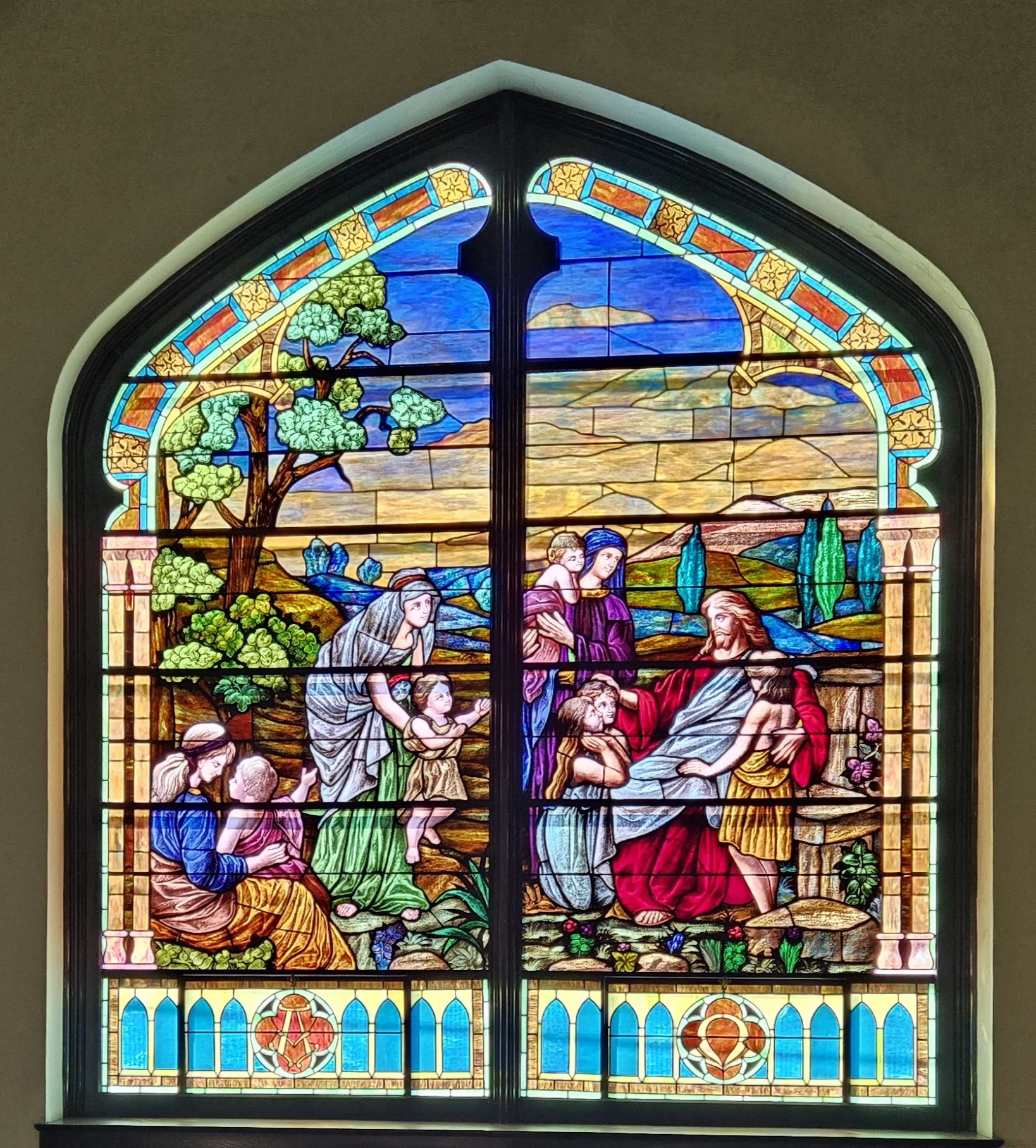 Colorful stained glass window depicting Jesus with children, surrounded by trees and landscape, in a church setting.