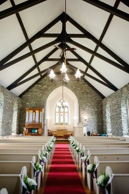 Interior of a small church with stone walls, wooden pews decorated with flowers, a red aisle runner, and a stained glass window at the front.