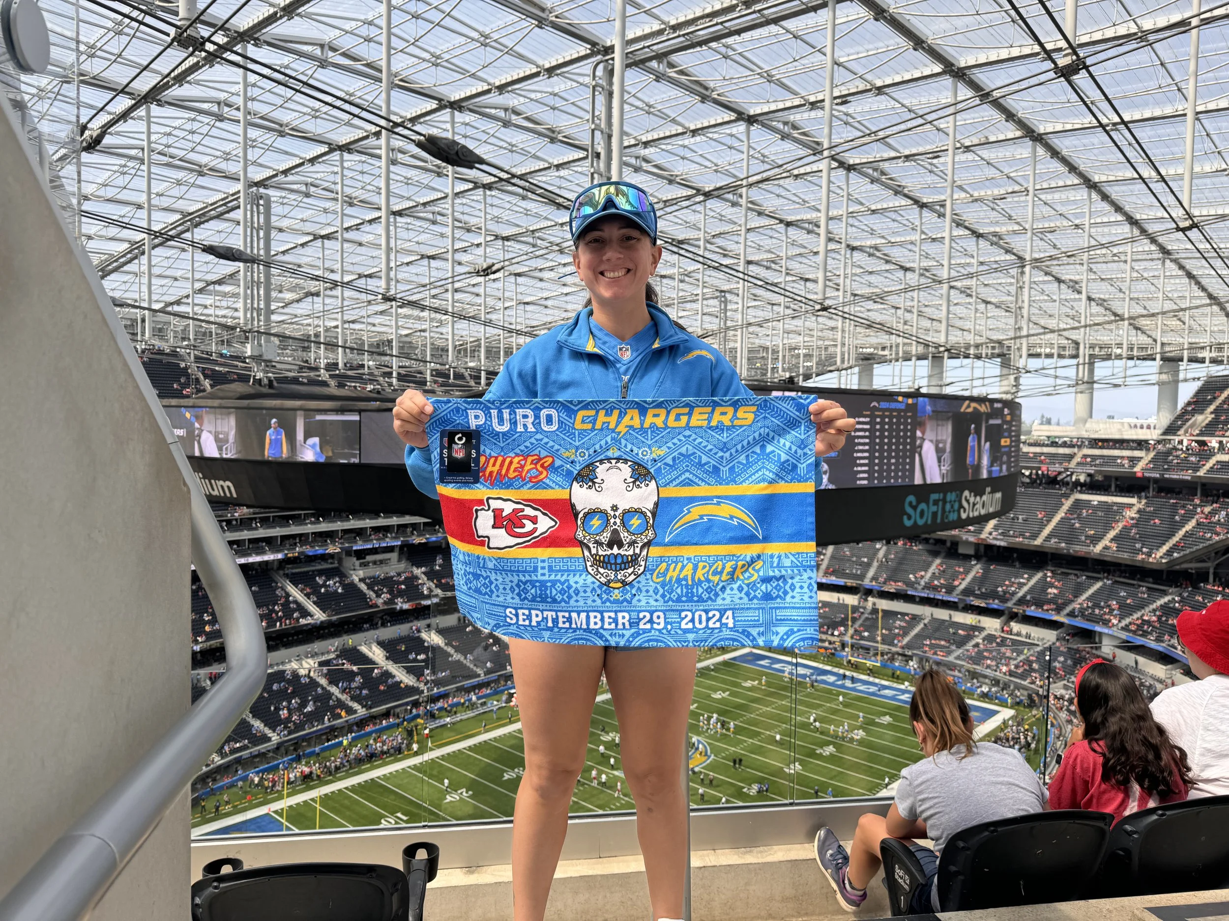 A woman at SoFi Stadium holding a Kansas City Chiefs-themed towel with the date September 29, 2024, smiling and wearing a blue Chargers jacket and sunglasses, with the stadium's seating and field in the background.