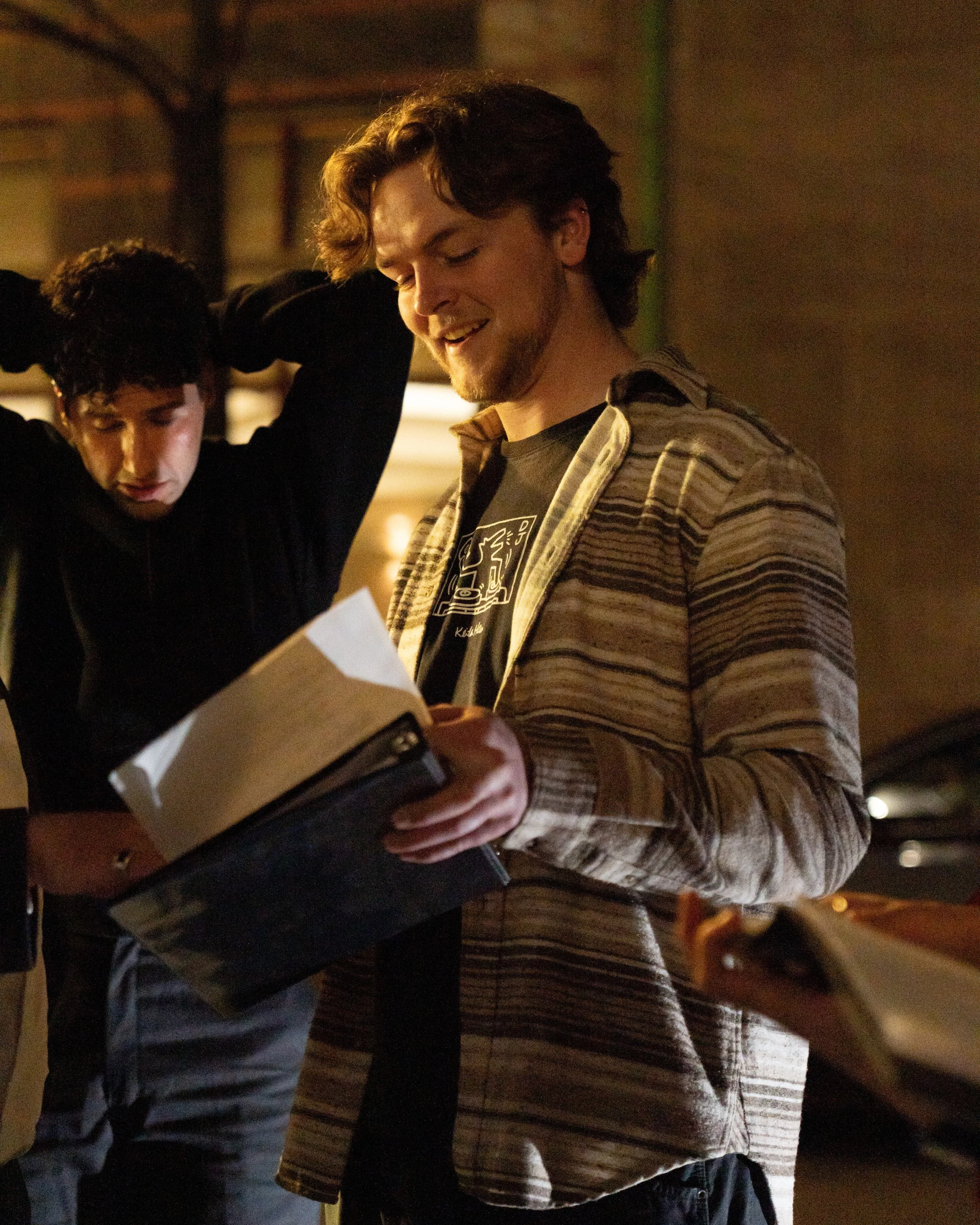 Two young adults, one with curly hair and the other with wavy hair, are standing close together in a dimly lit indoor setting. They appear to be smiling and engaged in reading or looking at something in a book or paper.