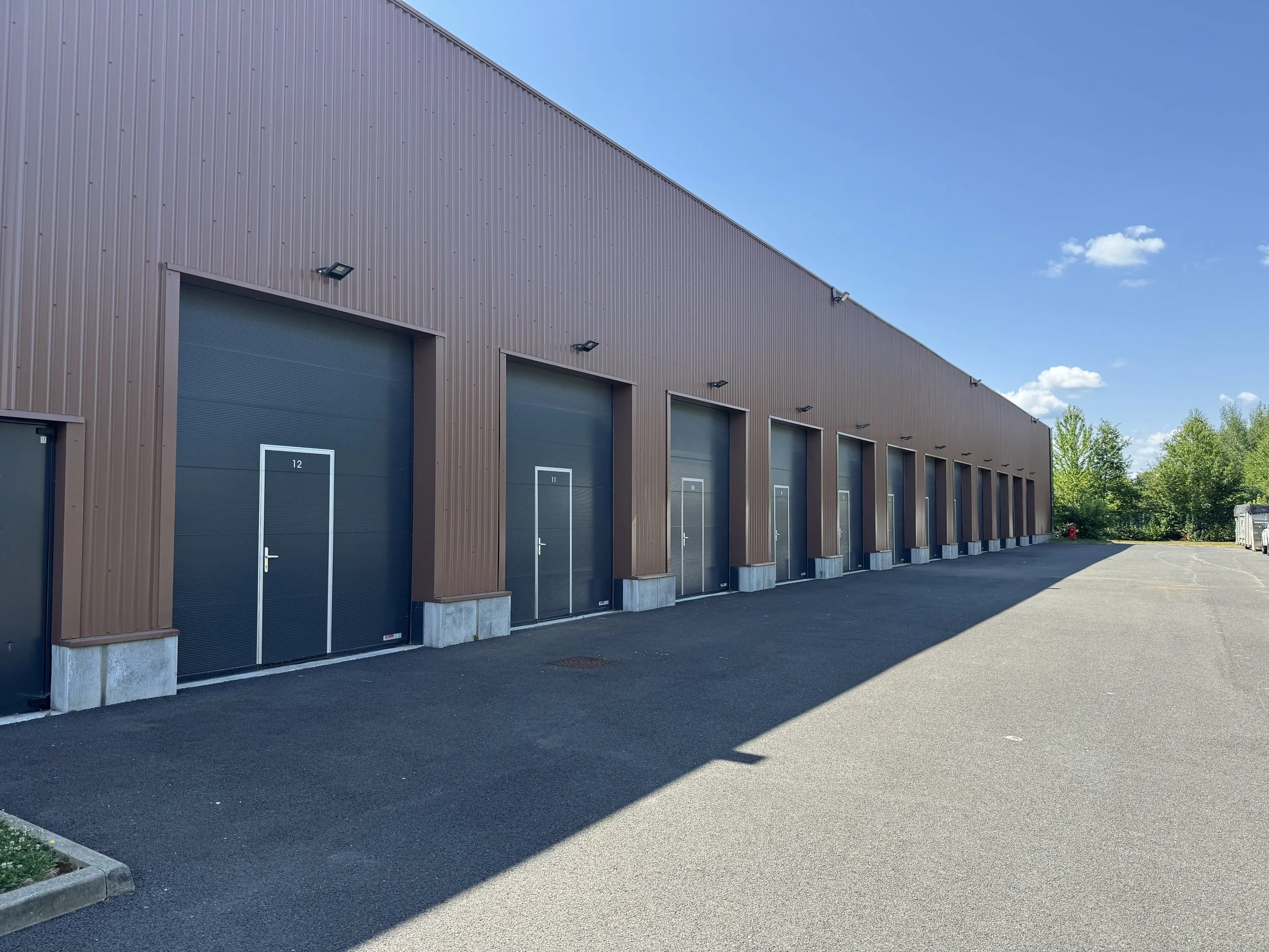 Une longue unité de stockage ou entrepôt avec plusieurs portes de garage numérotées 12, 11, etc., sous un ciel bleu avec quelques nuages, entourée d'arbres verts, situé à Serris.