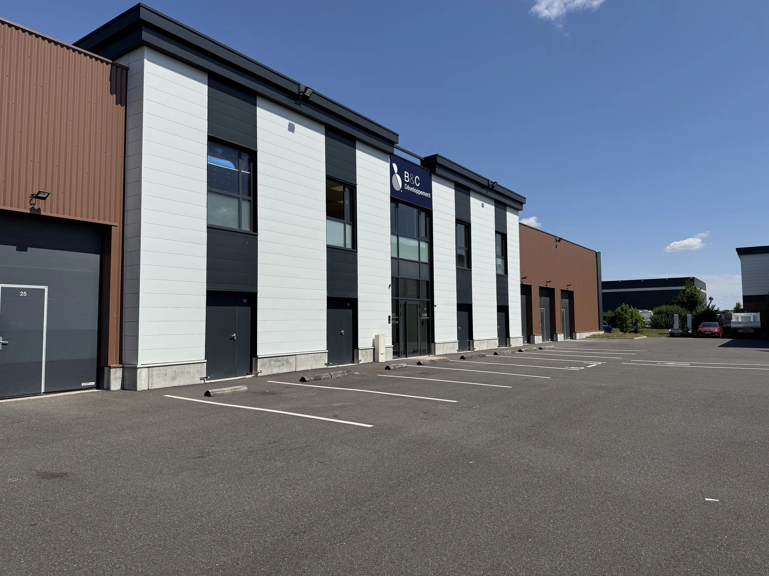 Bâtiment moderne avec un parking vide en premier plan, le bâtiment porte un panneau avec le texte 'B&C Développement', ciel bleu clair. Situé à Serris