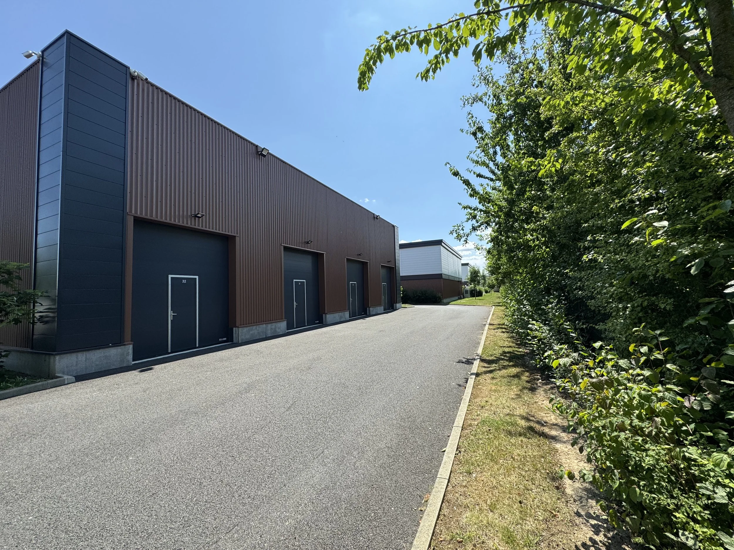 Façade d'un bâtiment industriel avec une allée en bitume et un trottoir, entouré d'arbustes et d'arbres, par une journée ensoleillée. A serris