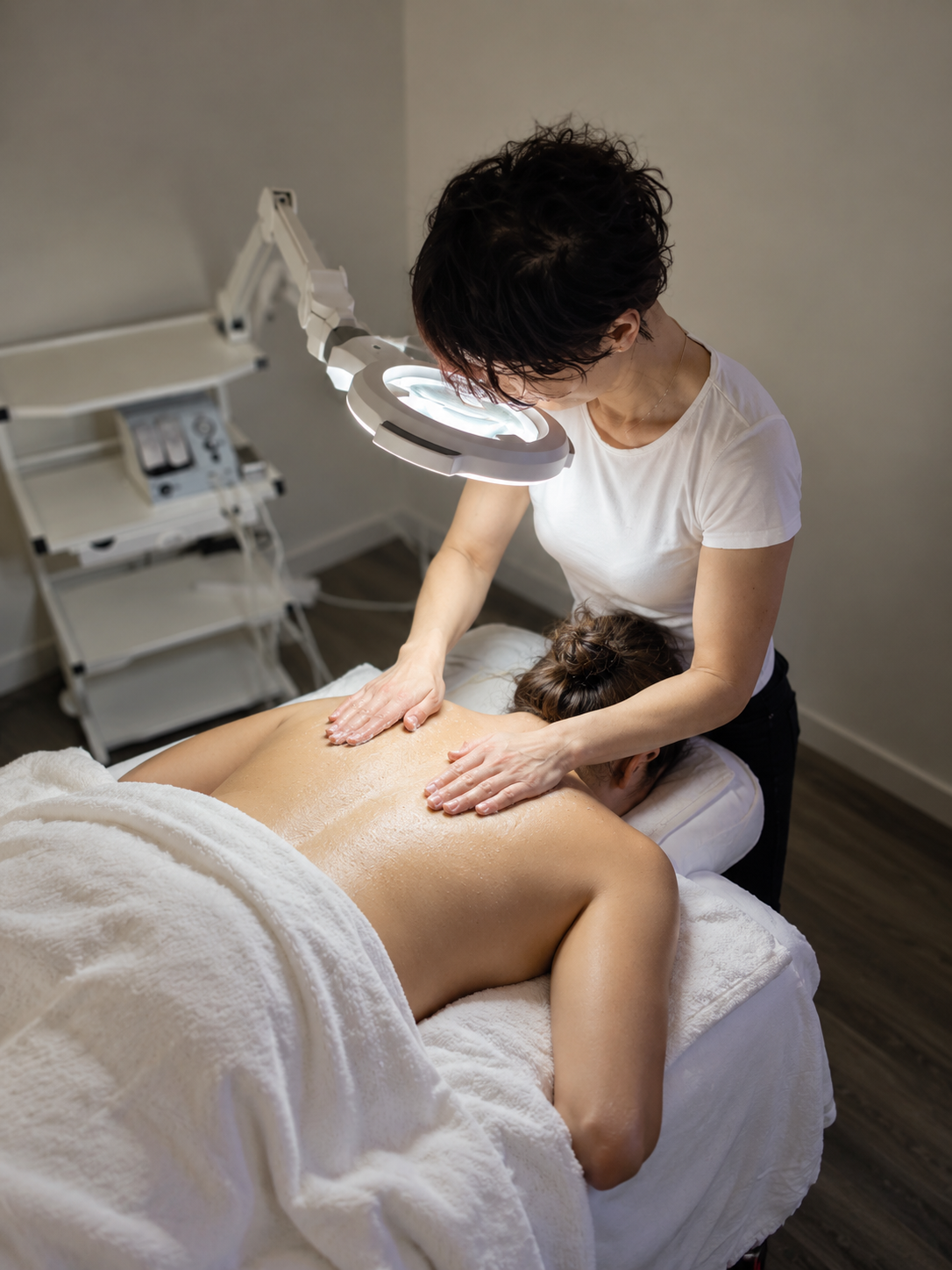 A woman receives a back facial treatment at Skincera Beauty from a therapist in a massage room, with a lamp overhead and medical equipment in the background.
