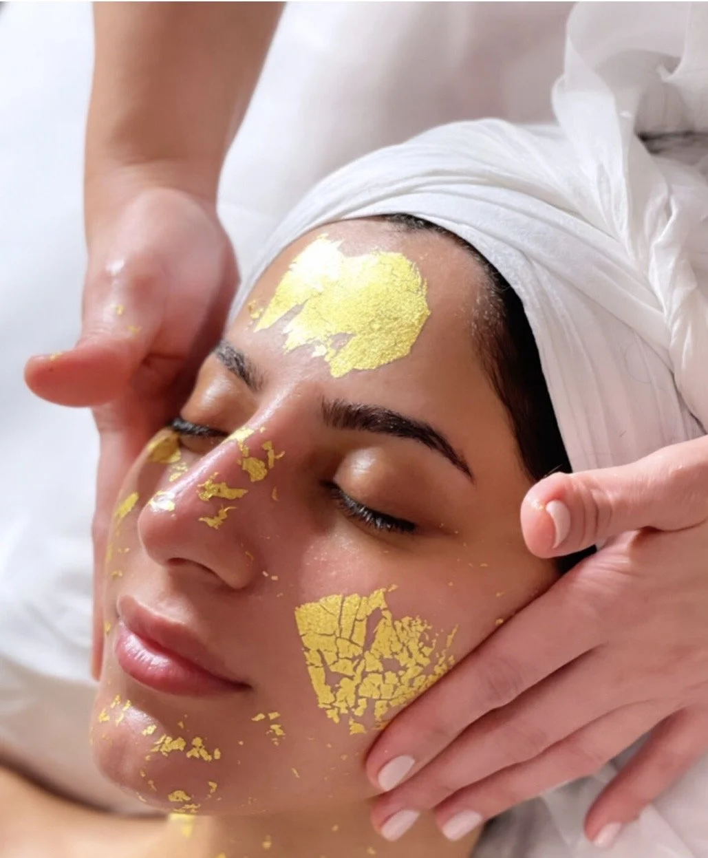 A woman is receiving a facial treatment with gold leaf applied to her face while lying with eyes closed, wearing a white head wrap.