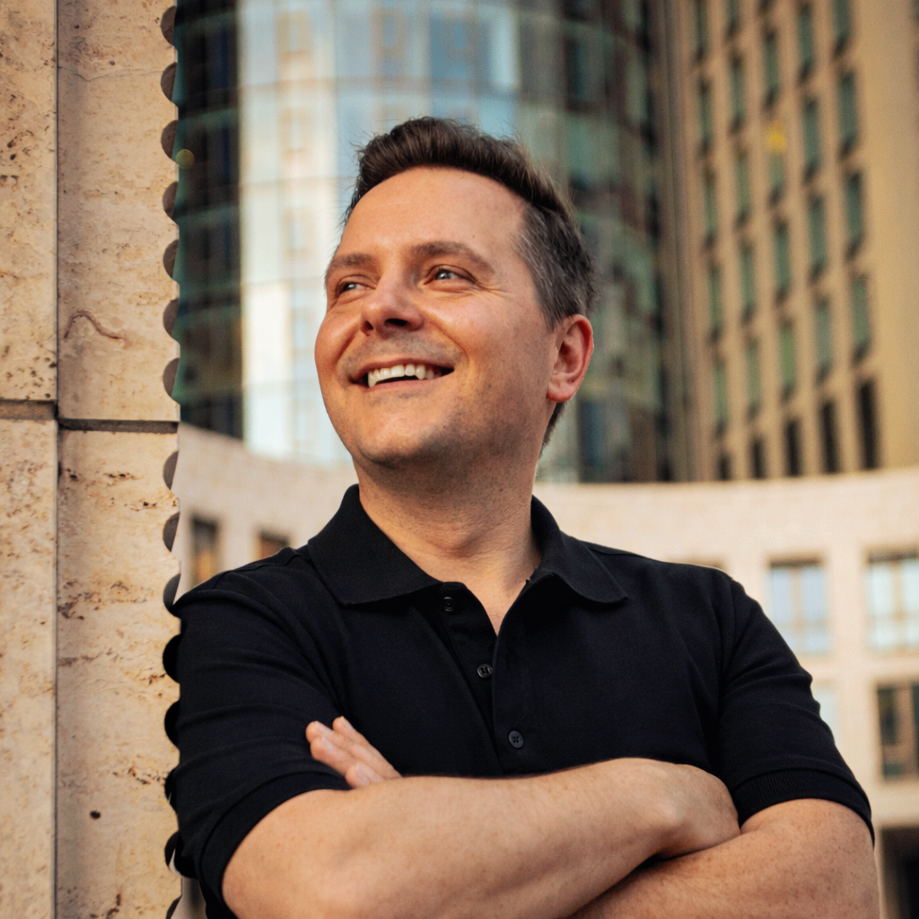 A smiling man with his arms crossed, wearing a black polo shirt, standing outdoors in an urban setting with tall buildings in the background.