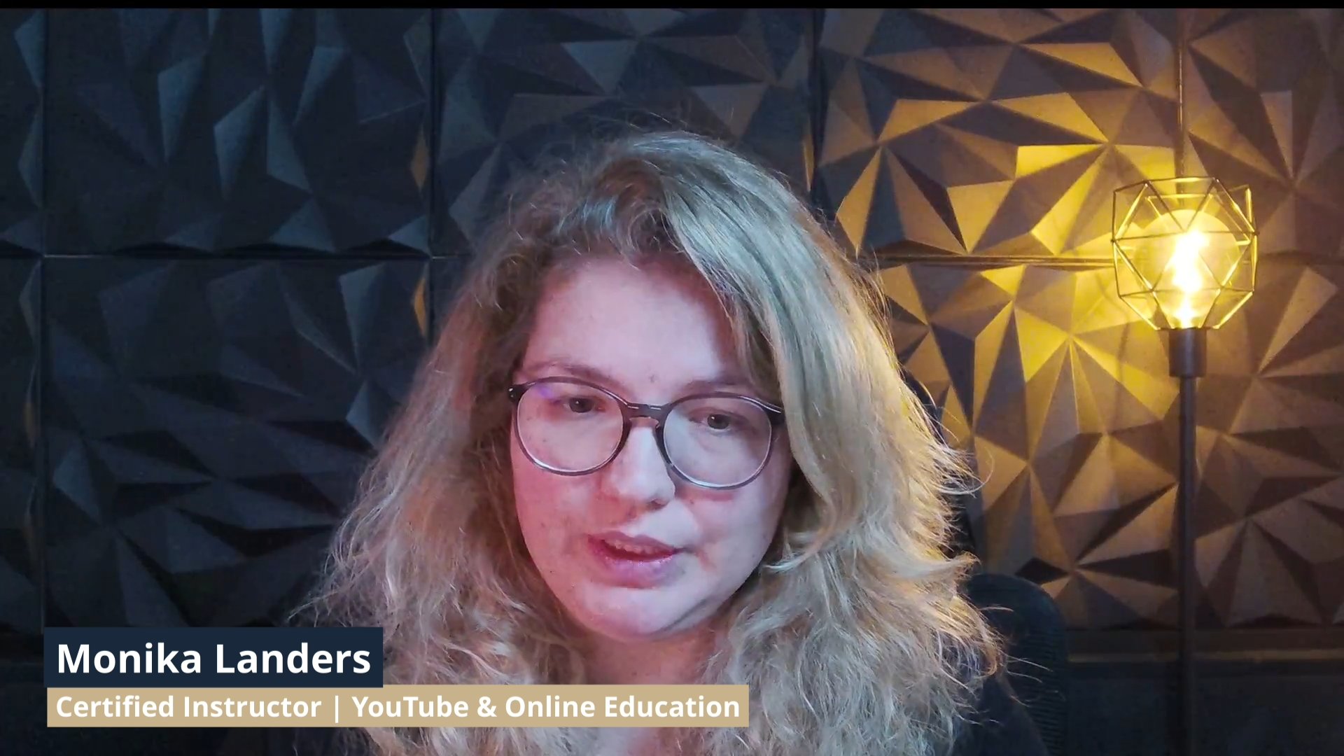 A woman with blonde curly hair and glasses speaking in front of a dark geometric textured wall with a warm yellow light fixture.