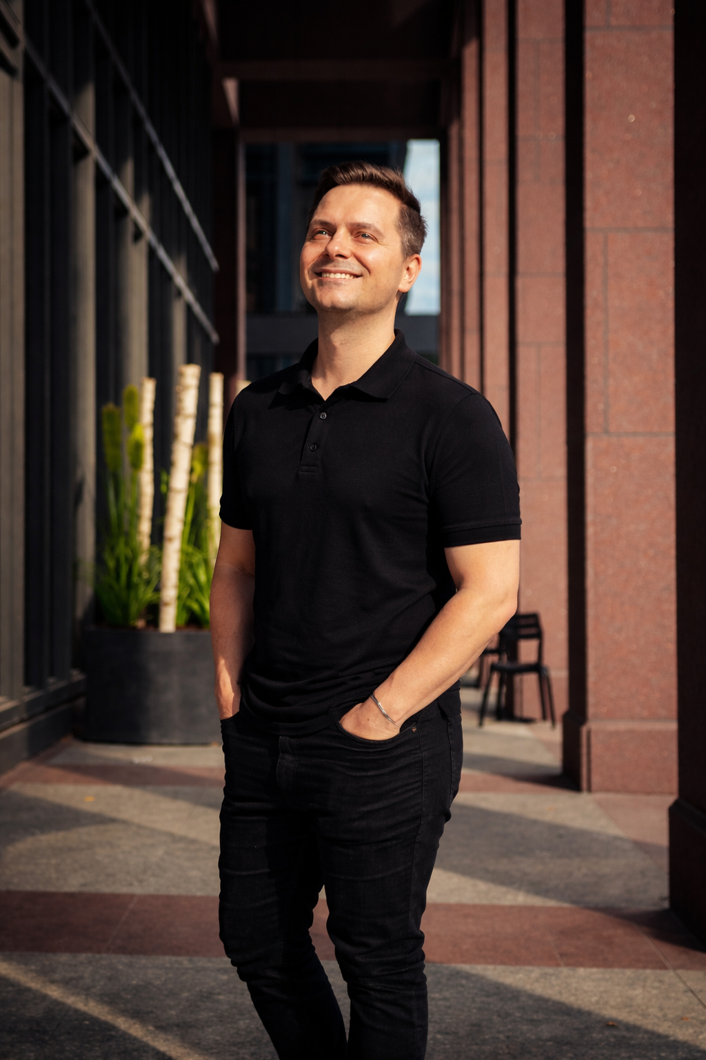 A smiling man wearing a black polo shirt and black jeans, standing outdoors with a modern building and green plants in the background.