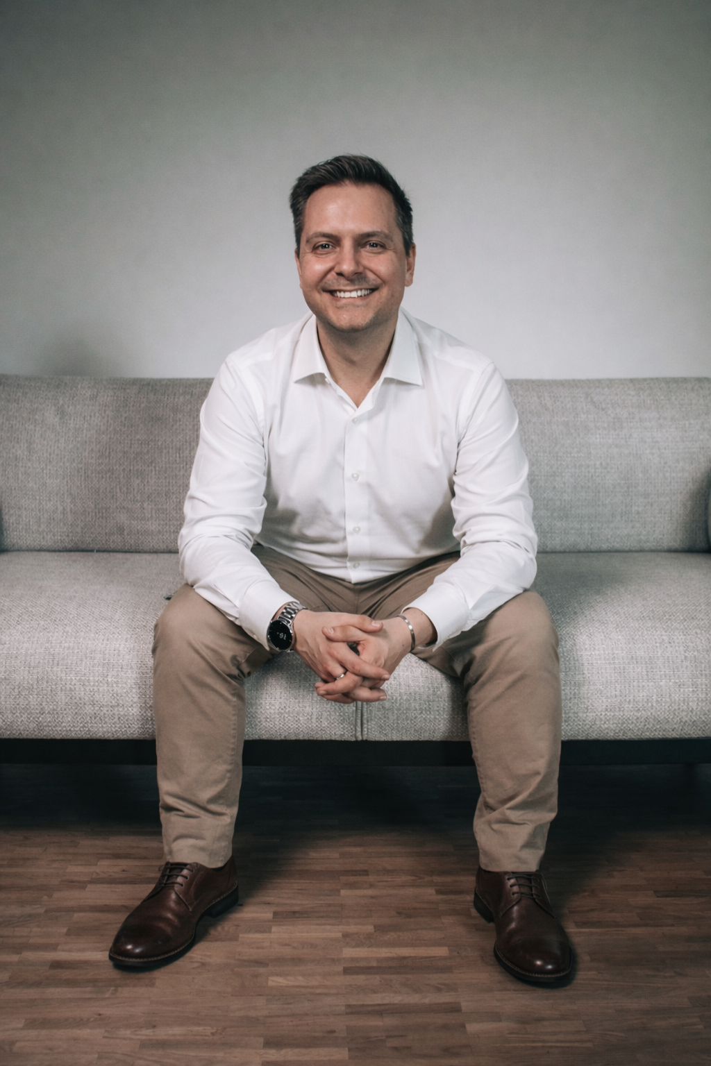 A man in a white shirt, beige pants, and brown shoes sitting on a gray sofa, smiling at the camera.