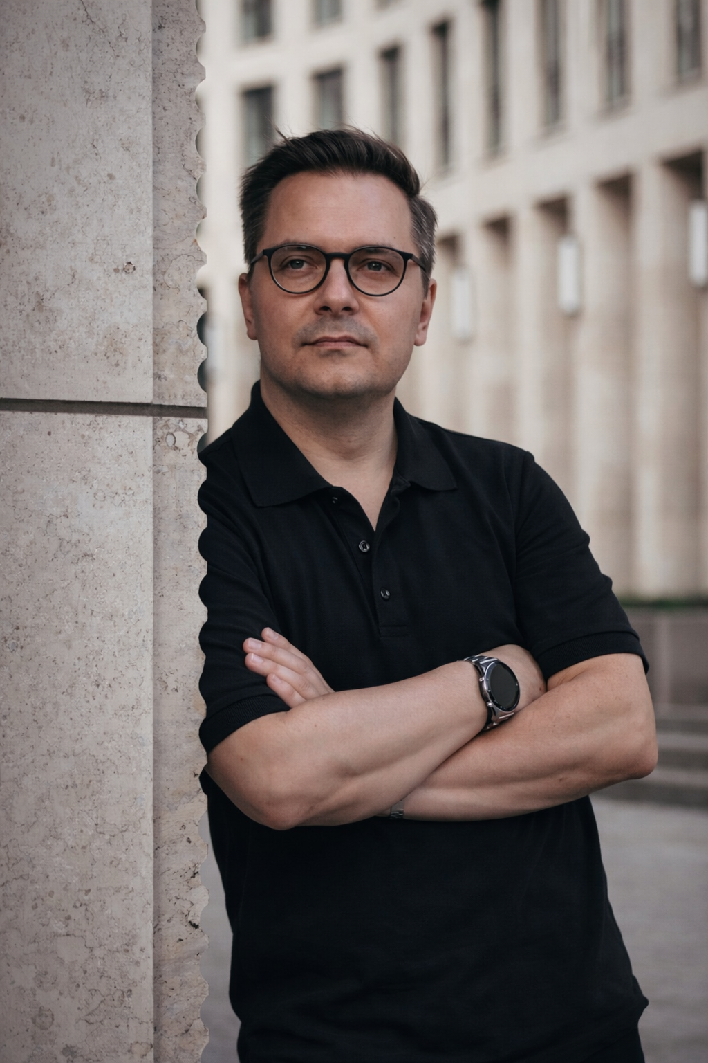 A man with dark hair and glasses leaning against a stone wall, with a serious expression, crosses his arms, wearing a black polo shirt, watch, and bracelet, in an urban setting with a multi-story building in the background.
