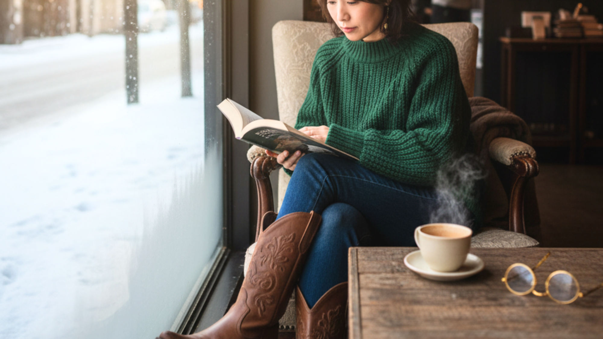 A Japanese women, 30 years old, wearing a green sweater, jeans, and brown cowboy boots reading a book in a cafe