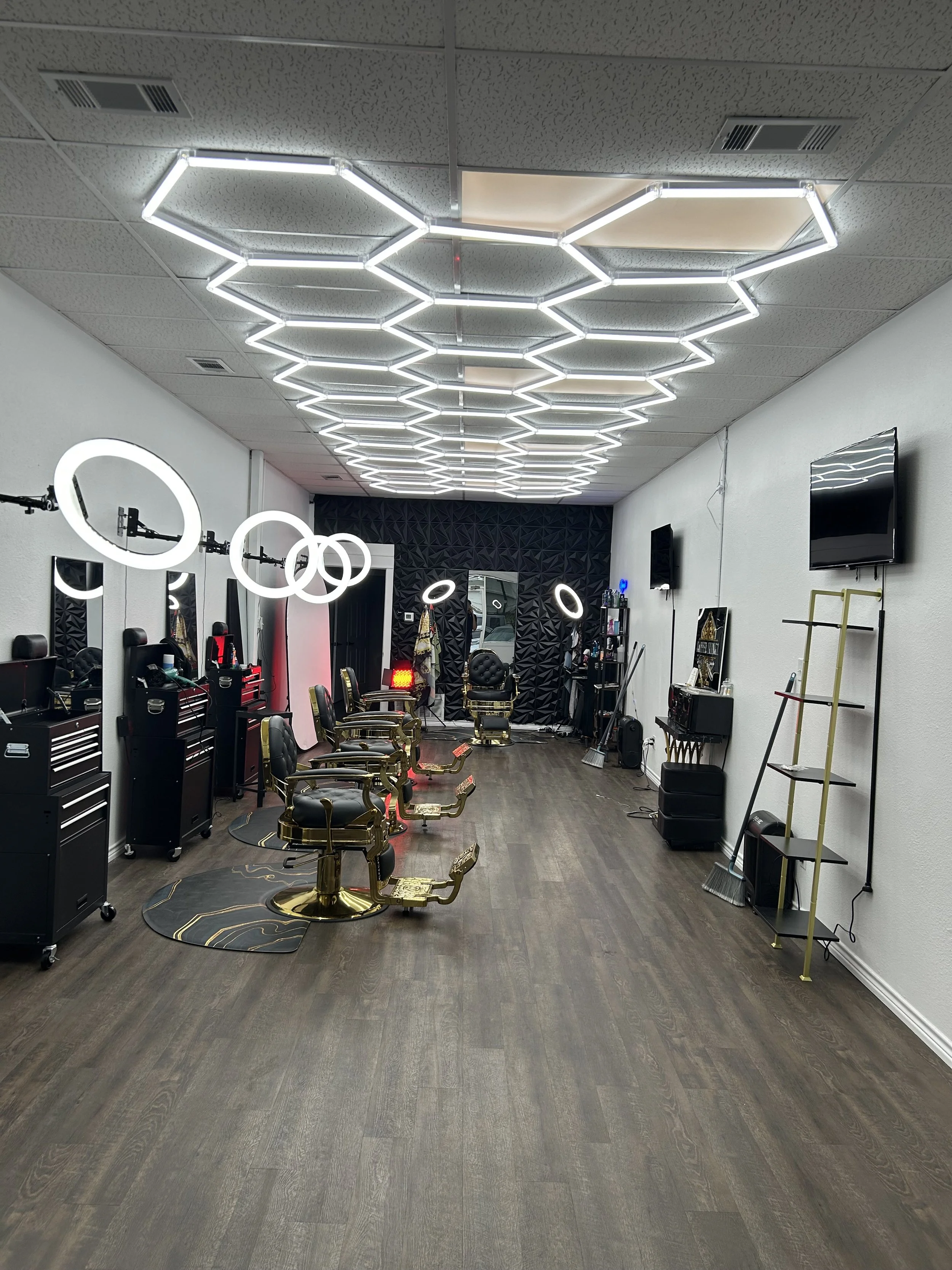 Empty modern barbershop with black and gold chairs, illuminated circular mirrors, LED ceiling lights, and barber tools on carts along the walls.