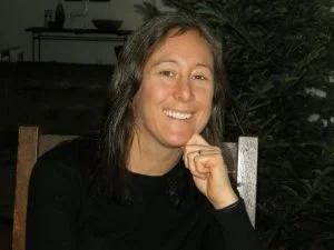 Smiling woman with dark hair sitting outdoors near a Christmas tree.