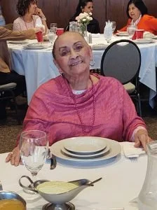 An elderly woman with a pink sweater sitting at a dining table in a restaurant, with a smile on her face, surrounded by other guests.
