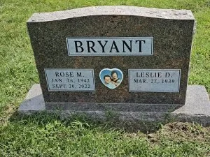Gravestone with the name Bryant, featuring two individual plaques with names Rose M. and Leslie D., their birth and death dates, and a heart-shaped photo of a couple in the center, set in grass.