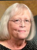 An elderly woman with shoulder-length gray hair, glasses, and a black dotted top, smiling against a wooden background.