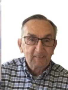 Close-up of an older man with glasses wearing a blue and white checkered shirt, smiling.