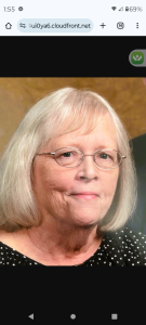 An elderly woman with shoulder-length gray hair, glasses, and a black dotted top, smiling against a wooden background.
