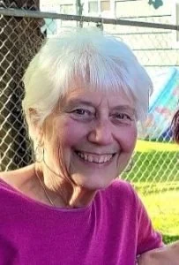 A smiling elderly woman with short white hair wearing a pink top outdoors near a chain-link fence, with a colorful playground in the background.