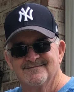 A man wearing a New York Yankees cap and sunglasses, smiling outdoors.