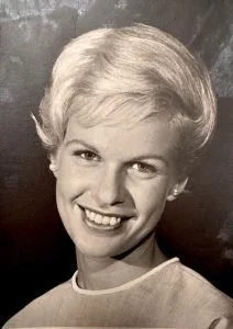 Black and white portrait of a smiling woman with short, curly, light-colored hair, wearing a light-colored top.