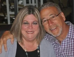 A smiling middle-aged couple posing together in a close-up shot, both with gray hair and glasses, in an indoor setting.