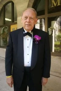 An older man in a tuxedo with a purple flower boutonniere, standing outdoors near a building.