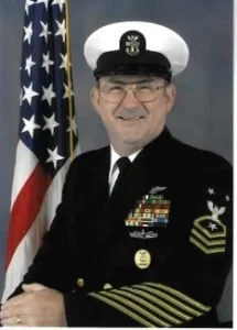 A smiling man in a U.S. Navy uniform, wearing glasses, with an American flag in the background.