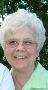 Close-up of an elderly woman with short white hair, glasses, and earrings, smiling outdoors with trees in the background.