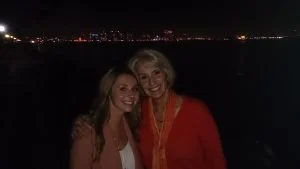 Two women smiling at night, standing outdoors with a city skyline in the background.