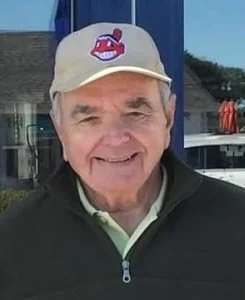An older man wearing a beige baseball cap with a Cleveland Indians logo, smiling outdoors with blue sky and buildings in the background.