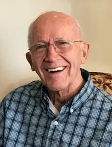 A smiling elderly man with glasses, wearing a blue plaid shirt, sitting indoors.