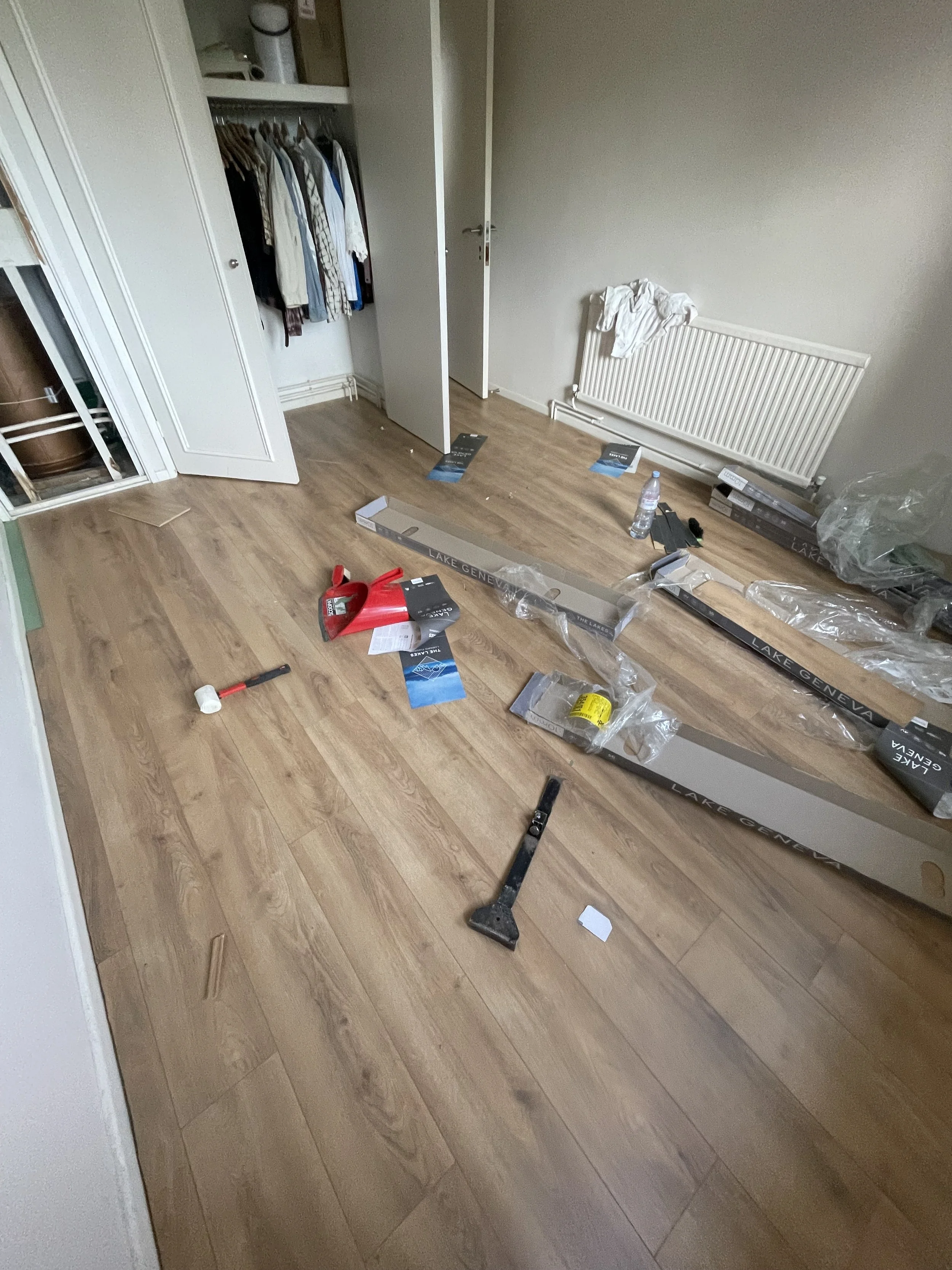 A room with wooden flooring, an open closet with clothes, a radiator, and various packaging materials and tools on the floor, indicating that furniture or fixtures are being assembled or installed.