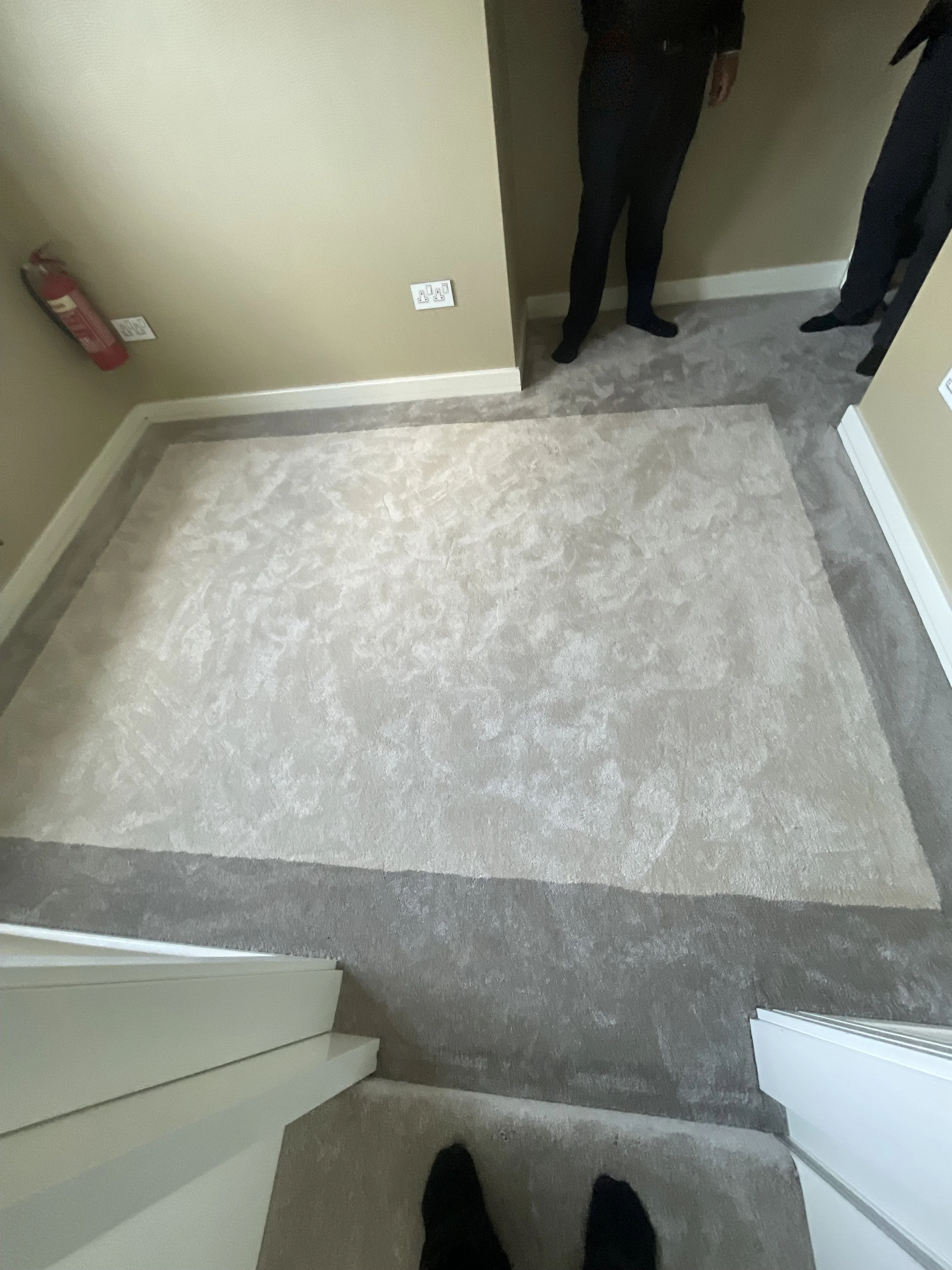 View from top of staircase showing a beige carpeted floor, a beige wall with electrical outlets, a fire extinguisher on the wall, and two people standing at the doorway, one wearing black pants and shoes.