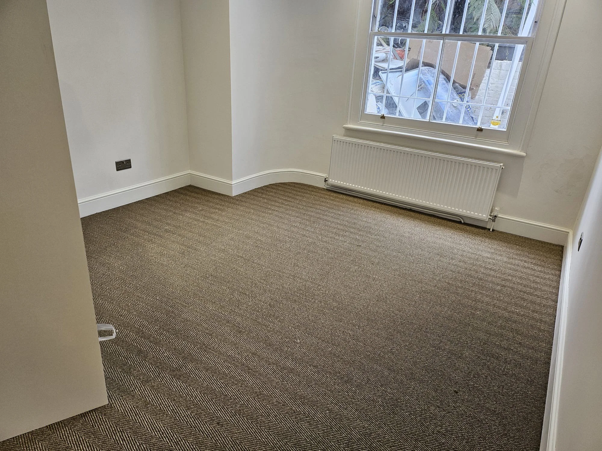 Empty room with beige carpet, white walls, a radiator under a window, and a view of outdoor clutter through the window.