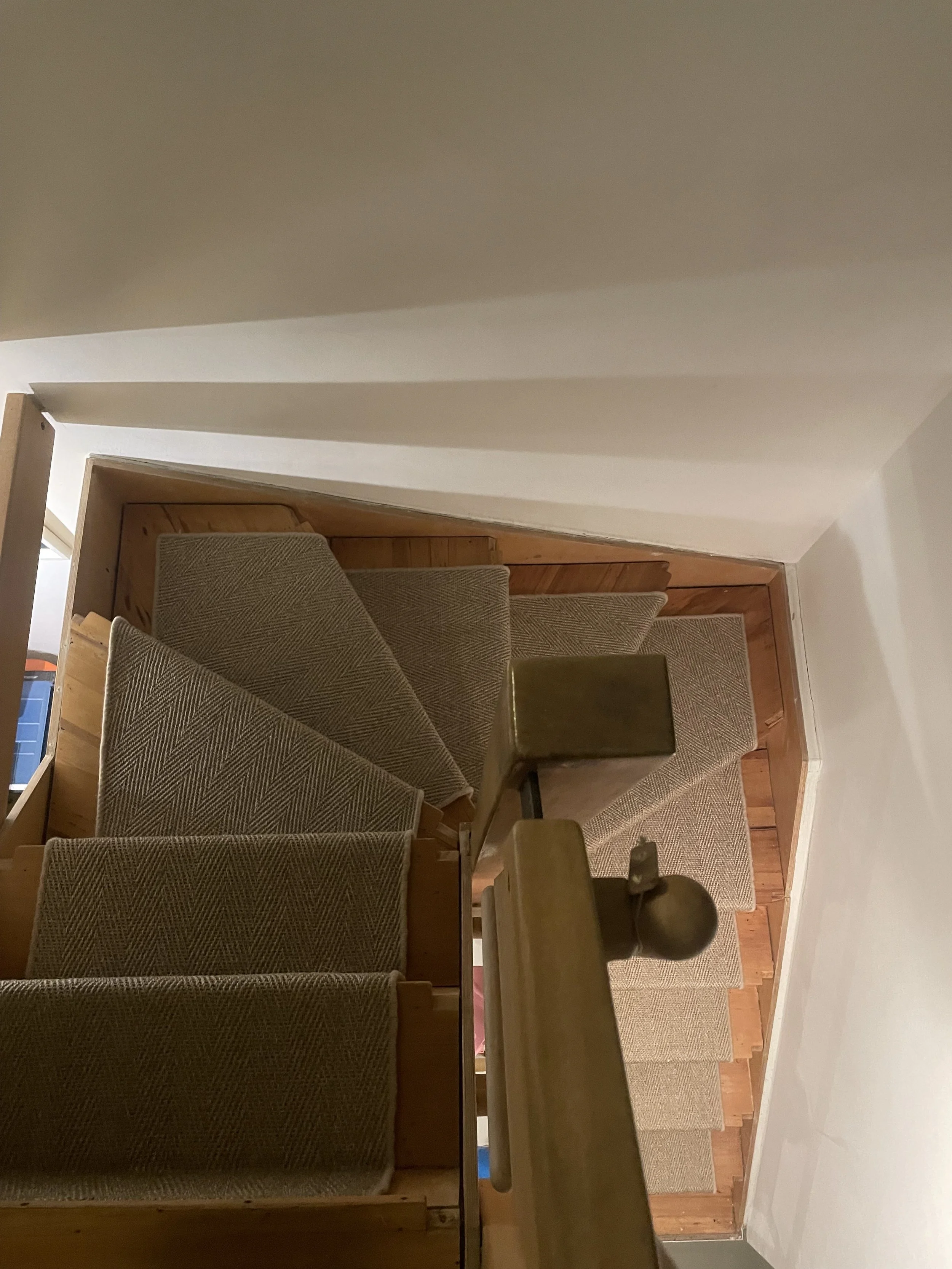 A staircase with carpeted steps viewed from above, with a wooden handrail and beige walls.